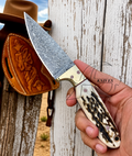 A hand holding a fixed-blade knife with a patterned steel blade and a carved bone handle, positioned in front of a rustic white wooden fence. A brown leather sheath and cowboy hat are visible in the background.