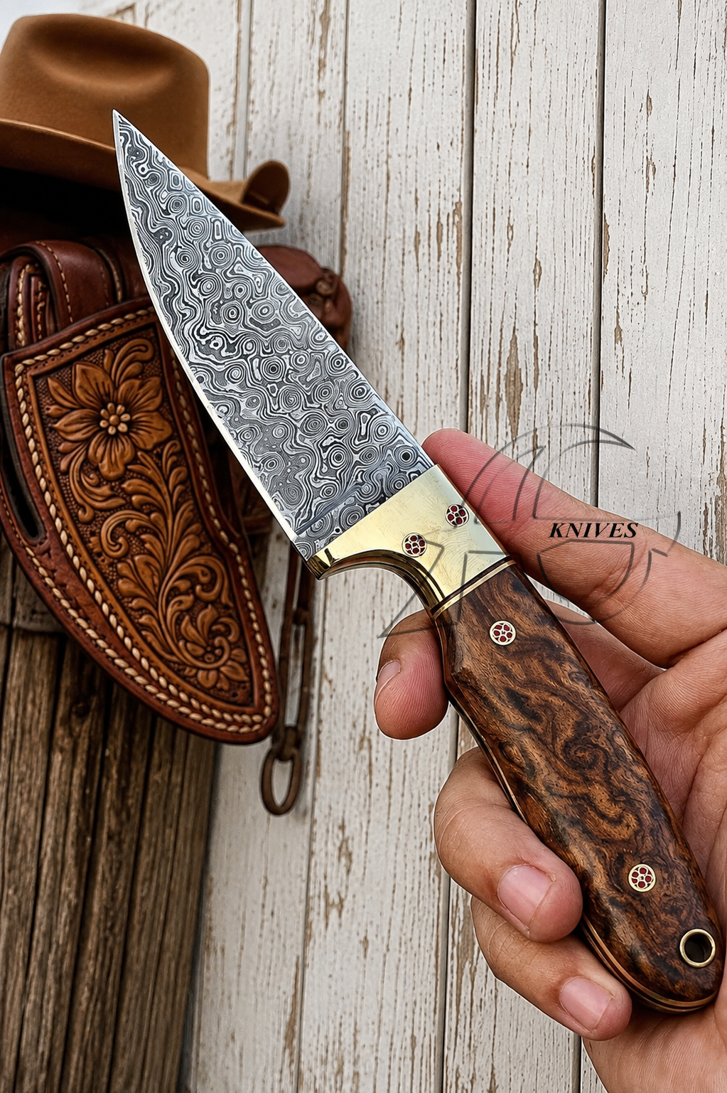 A hand holds a fixed-blade knife with a patterned steel blade, brass bolster, and dark wood handle in front of a brown leather sheath and cowboy hat on a rustic white wood plank background.