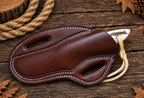 A brown leather knife sheath with white contrast stitching, shown on a rustic dark wood surface with a rope and autumn leaves in the background.