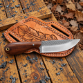 A fixed-blade knife with a dark wood handle and satin-finished steel blade rests on a tan leather sheath with an engraved feather design, placed on weathered wooden boards outdoors with scattered charcoal and dry leaves.